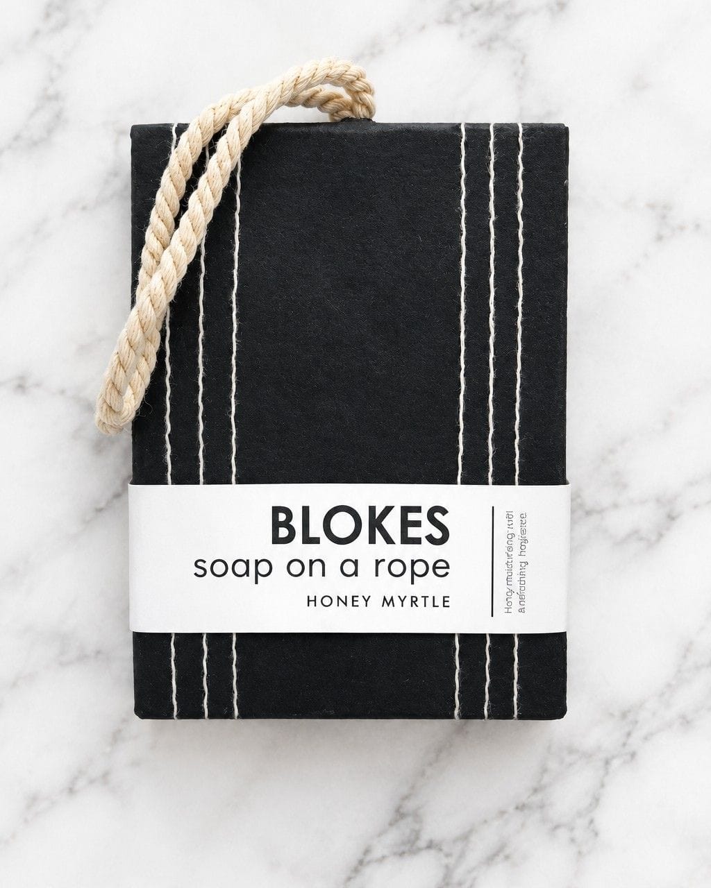 A packaged bar of soap called 'BLOKES soap on a rope' by Honey Myrtle, wrapped in a black cloth with white vertical stripes and a white paper band, featuring a natural rope handle, set on a white marble background.